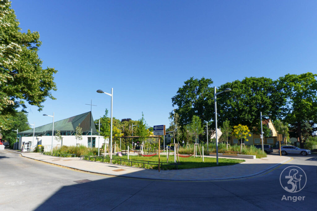 Der historische Dorfanger in Süßenbrunn mit einer gestalteten Grünfläche, Bäumen und Sitzgelegenheiten, im Hintergrund links ist die moderne Kirche Süßenbrunn unter blauem Himmel zu sehen.⁠