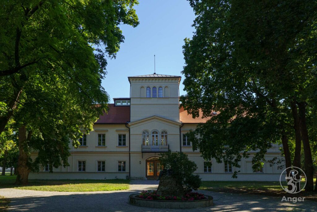 Die symmetrische Vorderansicht von Schloss Süßenbrunn in Wien mit dem charakteristischen Turmaufbau und Balkon, natürlich eingerahmt von großen, grünen Bäumen im Vordergrund.⁠