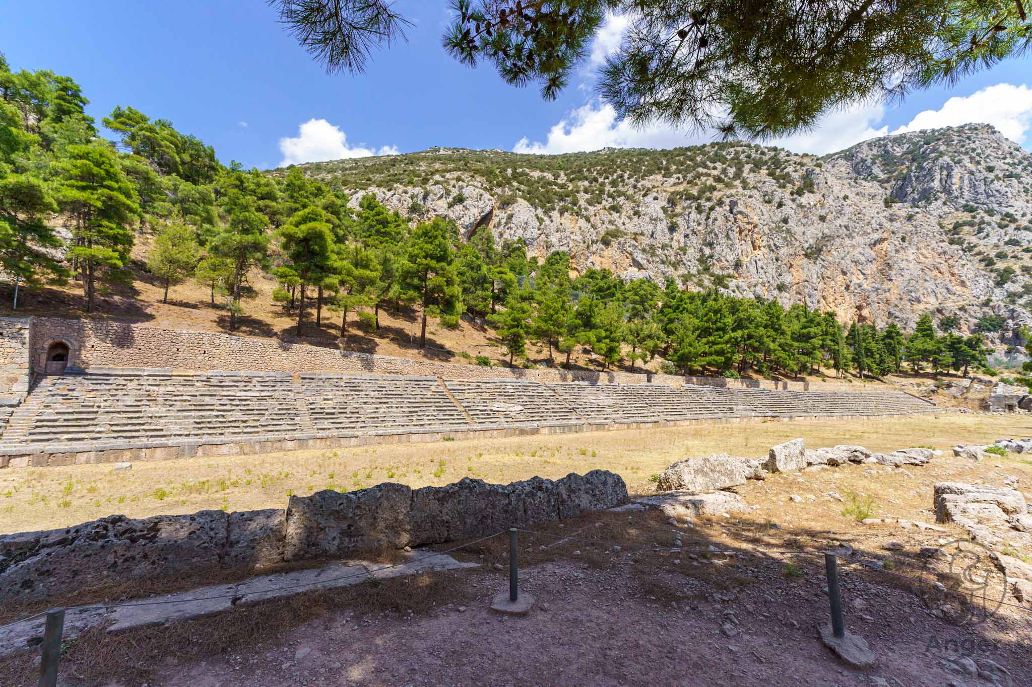 Langgestrecktes antikes Stadion in Delphi mit steinernen Tribünenrängen, umgeben von Nadelbäumen und felsigen Berghängen.
